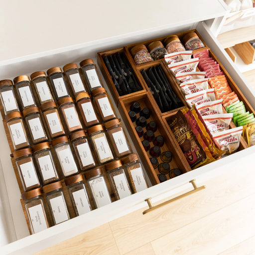 An open white drawer organized with glass spice jars, tea bags, and snack packets in wooden dividers. The drawer has a gold handle and is set against a light wood floor.