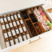 An open white drawer organized with glass spice jars, tea bags, and snack packets in wooden dividers. The drawer has a gold handle and is set against a light wood floor.