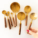 A set of brass measuring cups and spoons with wooden handles arranged on a white background, with a hand holding one of the larger cups.