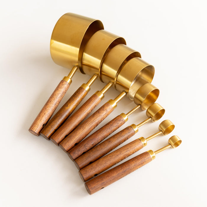 A set of nested measuring cups with shiny gold brass bodies and light brown wooden handles, arranged diagonally on a clean white background.