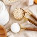 A styled flat-lay of kitchen ingredients and measuring tools on a beige linen cloth, including a glass jar of rolled oats, a small jar of pink salt, a white jar, a gold measuring cup filled with oats, and wooden-handled measuring spoons with one holding white sugar.